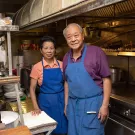 Two people stand in an old kitchen