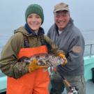 Woman smiling and holding a lingcod next to a man. The man is also smiling and holding a fishing pole.