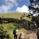 Group of individuals walk on a trail going up a hill. The sky is clear with clouds. 