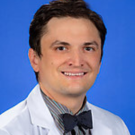 Close up headshot of a man smiling. He is wearing a white coat with a bowtie.