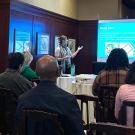 A group of people sits attentively in a conference room, watching a presenter who is standing next to a projection screen. The presenter, wearing a scarf and gesturing with one hand, is discussing a slide titled "Study Aims." The slide includes bullet points and colorful images. Attendees, seated in rows, focus on the presentation, some taking notes. A projector sits on a table in front of the screen, and framed artwork is visible on the wall behind the group.