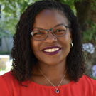 Close up headshot of a woman smiling outside. She is wearing a necklace and glasses.