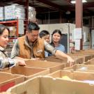 An Asian family of four, consisting of a father, mother and two children, stand at a table full of boxes as they help pack them with non-perishable items at a local Food Bank. They are each dressed casually and smiling at one another as they enjoy their time together helping their community.