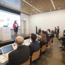 Woman in a bright red blazer and pencil skirt presents on a stage at the Manetti Shrem Museum in front of a crowd of people. The crowd is looking at her and most have laptops and notes with them.