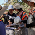 Five people wearing hats and orange safety vests are outdoors, looking at handheld equipment.
