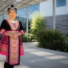Woman dressed in traditional Hmong clothing smiles outside in front of a building