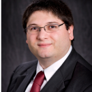 Close up headshot of a man wearing glasses and a suit in front of a plain background