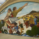 Large mural on the back wall of Woodland High School's theater building featuring  a woman with her back against a man. The man is facing away and points to the right. The woman holds a bowl and pours water to a field. They are behind large books, music notes and piano keys. Surrounding them are children holding arms and dancings. The mural is colorful and vibrant.