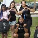 Three adults smile with two kids for a group photo. The kids are holding backpacks that say "Project Optimism Inc. Leader" and two women are smiling while making a circle with their hands. A man smiles for photo by squatting below the two women. He has two thumbs up facing outwards. 