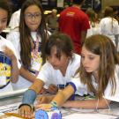 Young students wear white T Shirts with event logo and work on robotics projects indoors