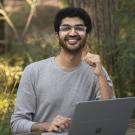 uc davis computer science student wearing glasses sits in front of a tree at a bench with his computer on the table