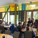A photo showing the community engagement summit at UC Davis. People are sitting at round tables and engaged in conversation. A few people are standing along the edge of the room. Colored windows in yellow, orange and blue are overhead.