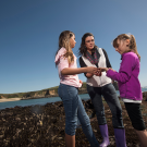 a woman and two young girls standing near a tidepool looking at something in their hands