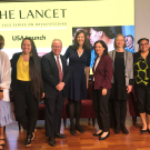 Nine individuals standing in a horizontal line smiling for a group photo. Each individual is wearing professional attire. They pose in front of a screen that reads “THE LANCET The 2023 Series on Breastfeeding USA Launch”