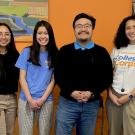 Four individuals stand side by side smiling for a close up group photo in front of a wall that has two artwork peeking on both the left and right side
