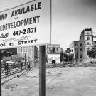 Black and white photo showing a large sign put up in front of a vacant lot. The sign reads &quot;Land Available for Redevelopment&quot; and gives a phone number and address. Buildings toward the back of the lot are undergoing various stages of demolition.