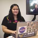 a young Latina woman looking at a laptop with lots of stickers, a screen in the background has a man standing in front of a forest.